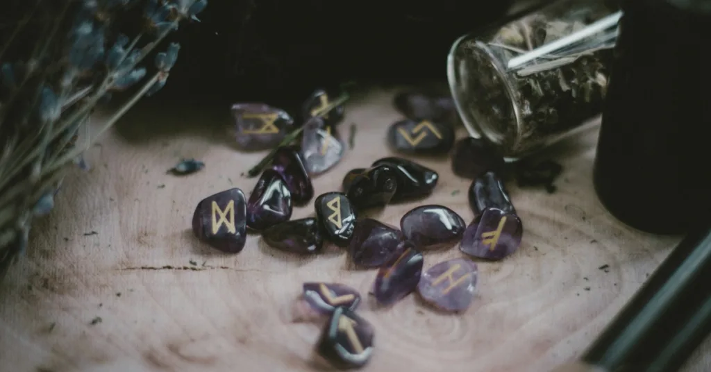 Elder Futhark runes engraved on amethyst stones lying on the table around magickal herbs.